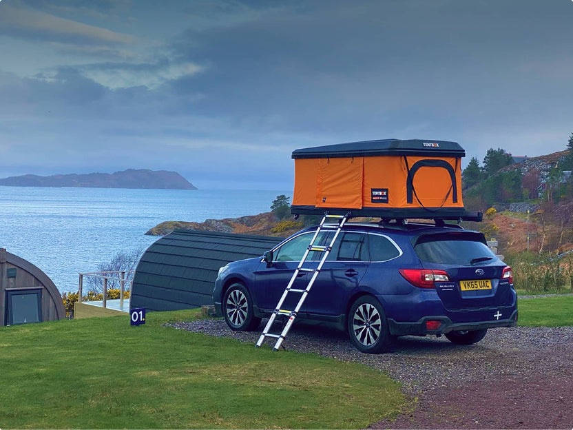Orange TentBox Classic set up on a vehicle in a coastal campsite, with ladder extended and sea in the background.