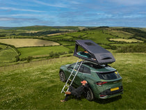 GO tent fully deployed on a car at a hilltop campsite with ladder extended; lush countryside in the background.

