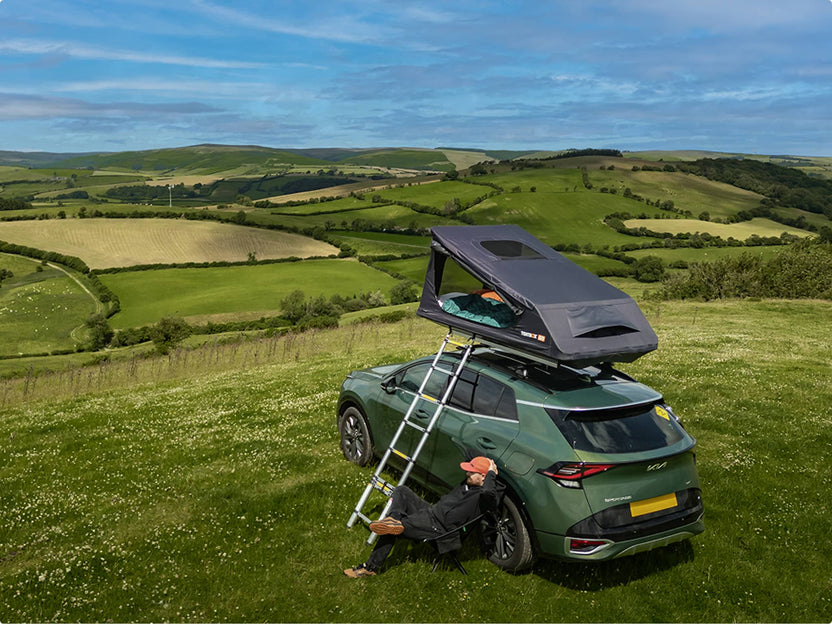 GO tent fully deployed on a car at a hilltop campsite with ladder extended; lush countryside in the background.

