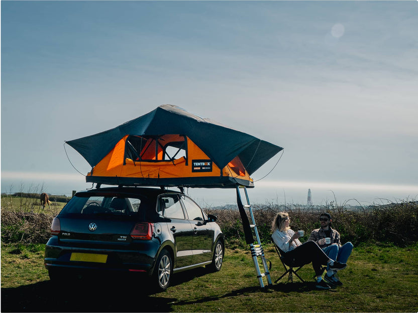 TentBox set up on a car in a coastal area, two people relaxing in camping chairs nearby