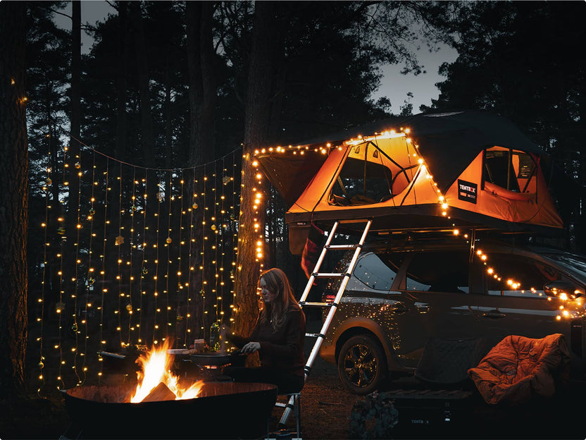 Nighttime scene with a TentBox lit up next to a firepit, fairy lights glowing in trees