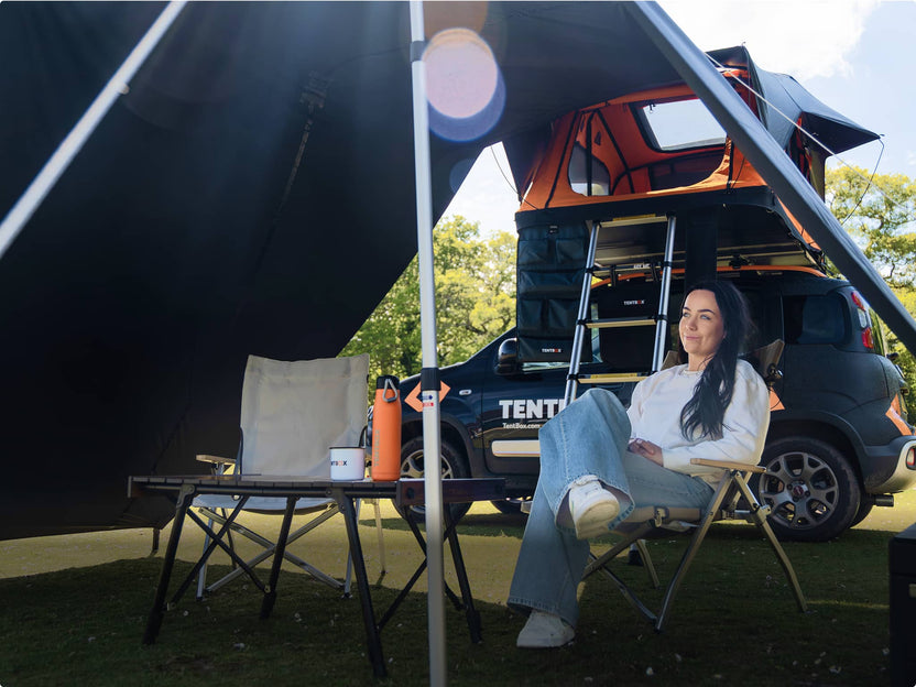 A woman relaxes under a TentBox awning beside a parked vehicle, sitting on a camping chair with a table, thermos, and mugs in front. The rooftop tent is extended, with the ladder visible