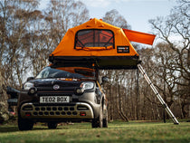 TentBox Lite 2.0 set up on top of a Fiat Panda in a wooded area. The tent is fully open, ladder extended to the ground, with trees and grass visible in the background