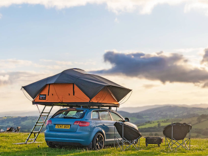TentBox Lite XL set up on a blue car in a scenic open area, with people relaxing on chairs nearby