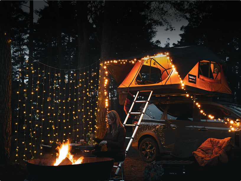 Cozy campfire scene with fairy lights and a TentBox glowing at night; ladder extended from vehicle to tent