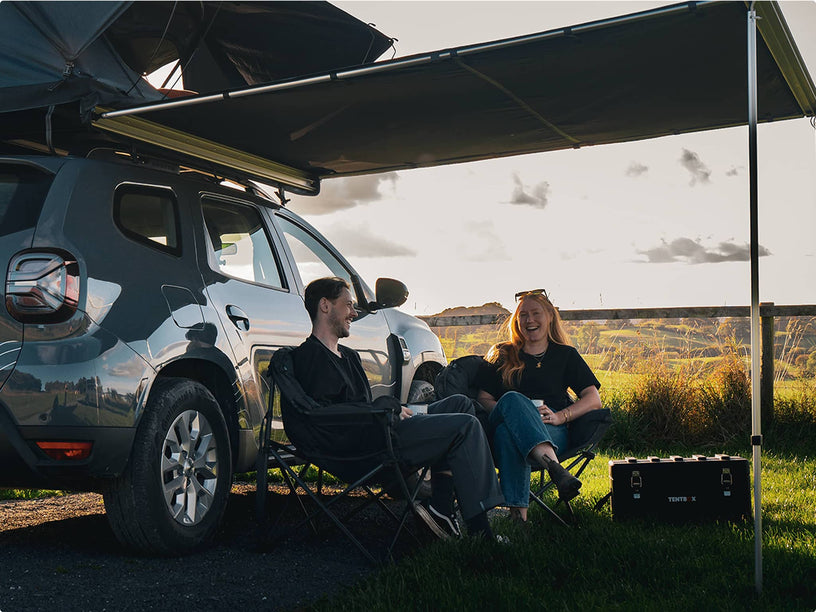 A man and woman sit comfortably under a TentBox awning attached to a silver SUV. They're laughing and enjoying a relaxed outdoor moment at sunset, with camping chairs and a cooler box beside them.